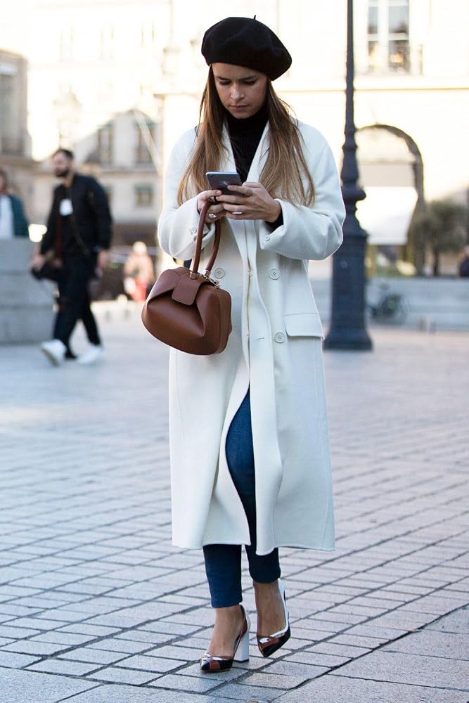 A woman walking outdoors on a paved street wearing a long white coat, black french beret, black top, blue jeans, and brown heels, while holding a brown leather handbag and looking at her phone.